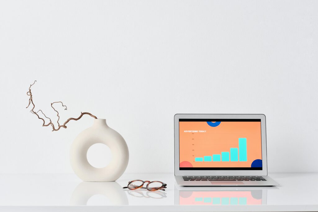 A minimalist workspace with a laptop, ceramic vase, and eyeglasses on a white desk.